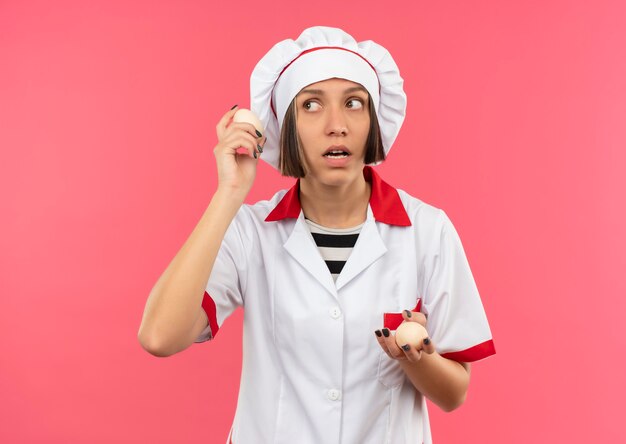 Giovane cuoco femminile premuroso in uniforme del cuoco unico che tiene le uova e che esamina il lato isolato sulla parete rosa