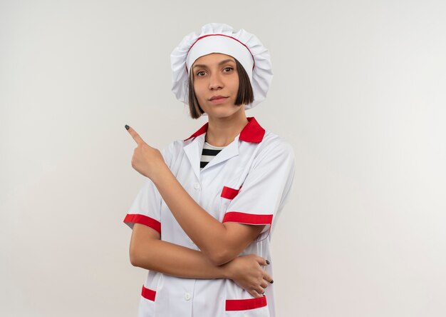 Giovane cuoco femminile sicuro in uniforme del cuoco unico che indica al lato isolato su bianco con lo spazio della copia