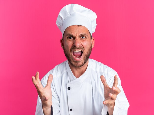 Giovane cuoco maschio caucasico arrabbiato in uniforme da chef e berretto che tiene le mani in aria urlando