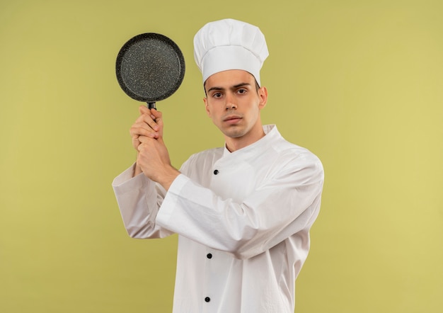 giovane cuoco maschio che indossa l'uniforme del cuoco unico che tiene la padella intorno alla spalla sulla parete verde isolata