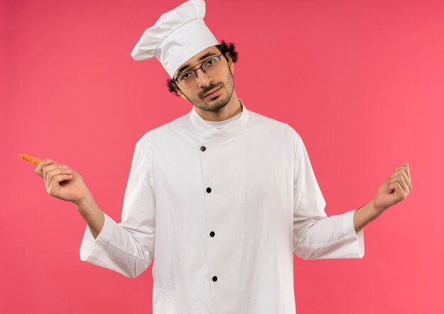 giovane cuoco maschio che indossa l'uniforme dello chef e bicchieri che tengono la carota e allarga i pugni