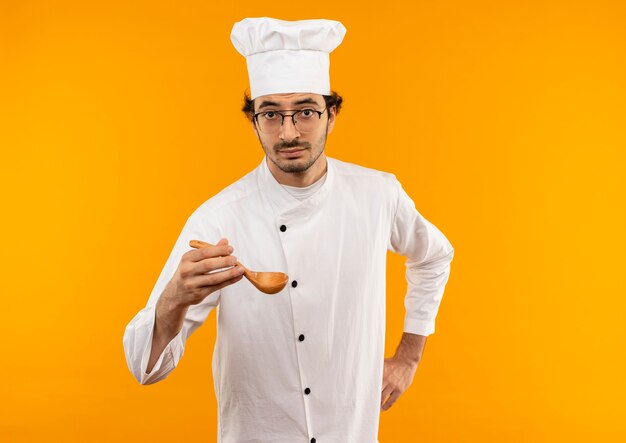 giovane cuoco maschio che indossa l'uniforme dello chef e bicchieri tenendo il cucchiaio mettendo la mano sul fianco