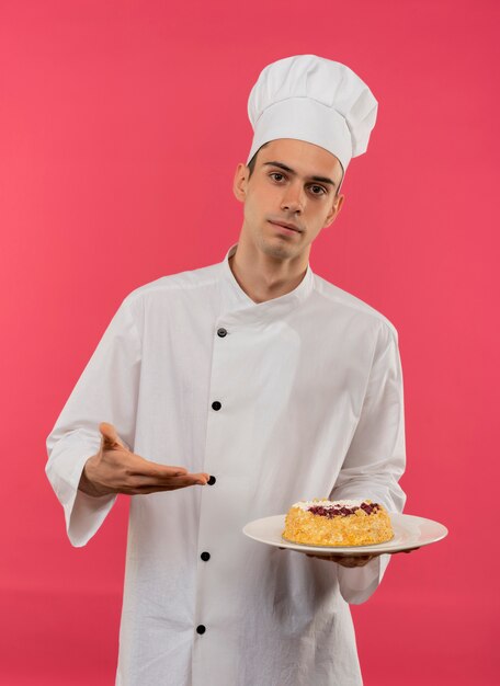 giovane cuoco maschio che indossa la tenuta uniforme dello chef e punti con la torta di mano sul piatto sul muro rosa isolato