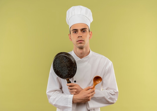 giovane cuoco maschio che indossa uniforme da chef incrocio padella e cucchiaio in mano sul muro verde isolato con spazio di copia