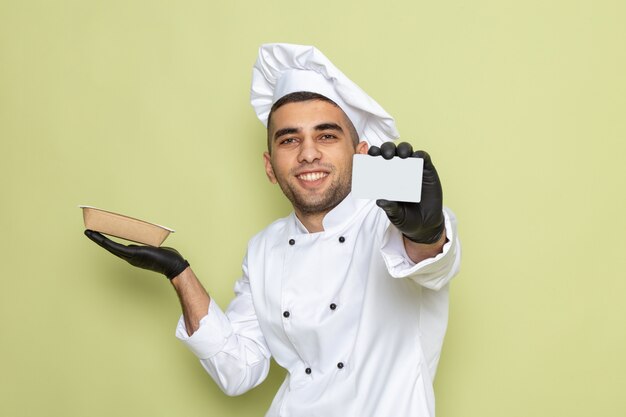 Giovane cuoco maschio di vista frontale in vestito bianco del cuoco che porta i guanti scuri che tengono alimento del pacchetto sul verde