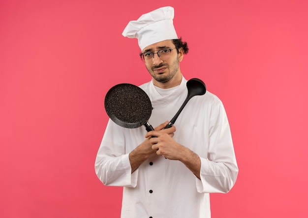 Giovane cuoco maschio dispiaciuto che indossa l'uniforme del cuoco unico e vetri che tengono e che attraversano la padella e la spatola sul rosa