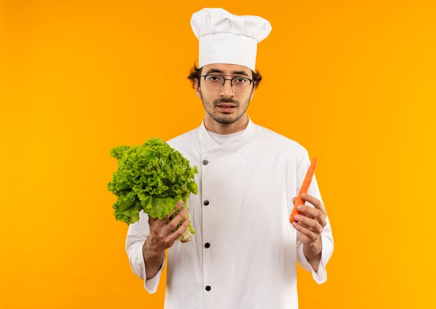 Giovane cuoco maschio dispiaciuto che indossa l'uniforme del cuoco unico e vetri che tengono insalata e carota isolate sulla parete gialla