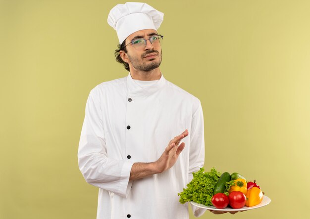 Giovane cuoco maschio dispiaciuto che indossa l'uniforme dello chef e bicchieri tenendo le verdure sul piatto e mostrando il gesto di arresto