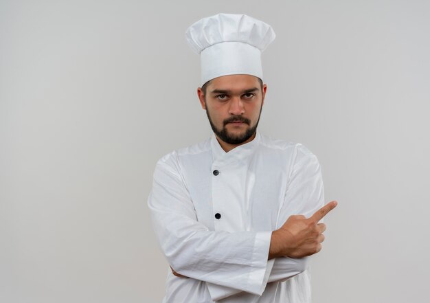 Giovane cuoco maschio in uniforme del cuoco unico che sta con la postura chiusa e che indica al lato