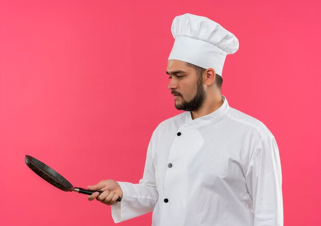 Giovane cuoco maschio in uniforme del cuoco unico che tiene e che esamina la padella isolata sullo spazio rosa