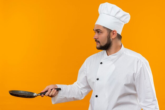 Giovane cuoco maschio in uniforme del cuoco unico che tiene la padella e guardando il lato isolato sullo spazio arancione