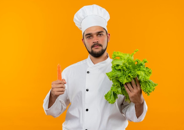 Giovane cuoco maschio in uniforme del cuoco unico che tiene lattuga e carota che sembrano isolati sullo spazio arancione