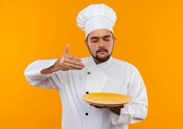 Giovane cuoco maschio nel piatto della tenuta dell'uniforme del cuoco unico e annusando con la mano sull'aria e gli occhi chiusi isolati sullo spazio arancione