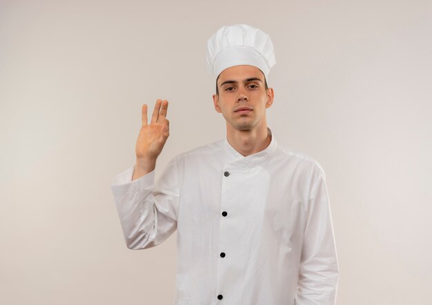 Giovane cuoco maschio sicuro che indossa l'uniforme del cuoco unico che mostra okey gesto sulla parete bianca isolata con lo spazio della copia