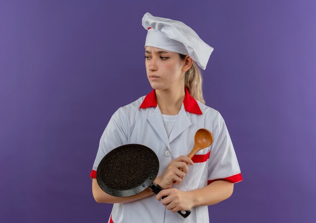 Giovane cuoco poco soddisfatto in uniforme del cuoco unico che tiene la padella e il cucchiaio che esaminano lato isolato sullo spazio viola