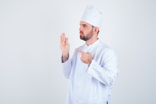 Giovane cuoco unico maschio che indica alla sigaretta in uniforme bianca e che sembra premuroso. vista frontale.