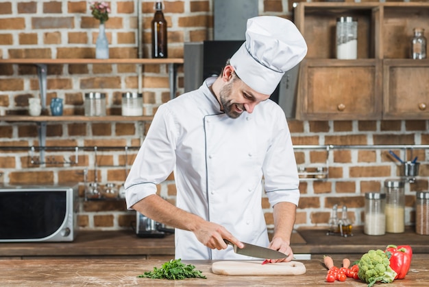 Giovane cuoco unico maschio felice che taglia peperoncino rosso rosso con il coltello sul contatore di cucina