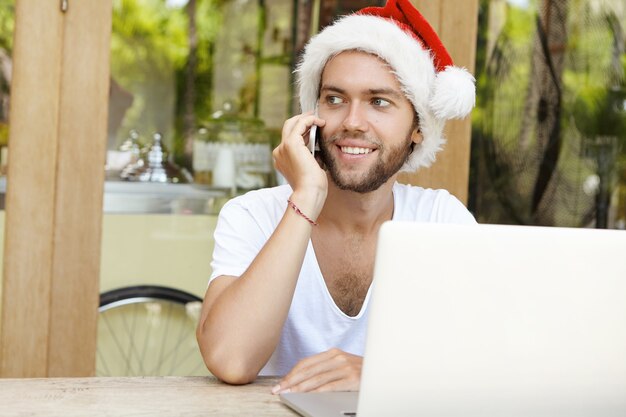 Giovane di successo e felice in cappello rosso con pelliccia bianca che trascorre le vacanze di Natale nel paese caldo, avendo conversazione telefonica con il suo amico mentre utilizza il wi-fi gratuito su un computer portatile generico