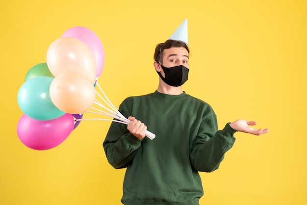 Giovane di vista frontale con cappuccio del partito e palloncini colorati che aprono la mano in piedi sul giallo