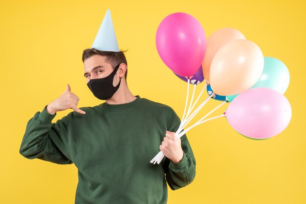 Giovane di vista frontale con cappuccio del partito e palloncini colorati che fanno chiamarmi segno del telefono in piedi sul giallo