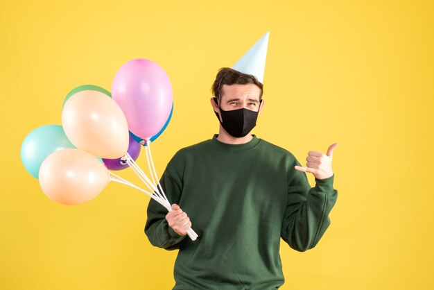 Giovane di vista frontale con cappuccio del partito e palloncini colorati che fanno chiamarmi segno di telefono su giallo
