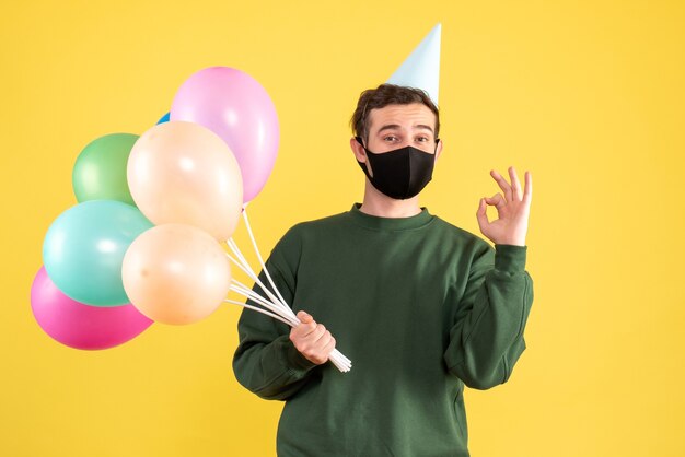 Giovane di vista frontale con cappuccio del partito e palloncini colorati che fanno segno di okey in piedi sul giallo