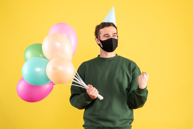 Giovane di vista frontale con cappuccio del partito e palloncini colorati che mostrano il gesto vincente sul giallo