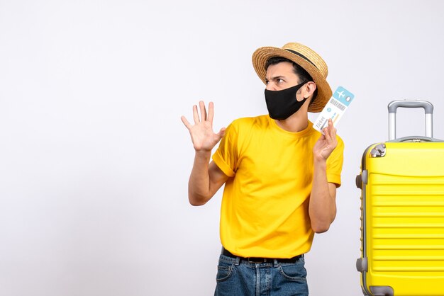 Giovane di vista frontale con il cappello di paglia che sta vicino al biglietto aereo giallo della tenuta della valigia