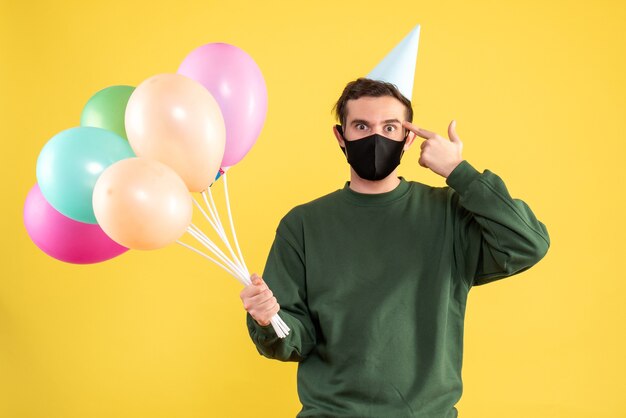 Giovane di vista frontale con il cappuccio del partito e palloncini colorati che pronunciano la pistola del dito alla sua tempia su fondo giallo