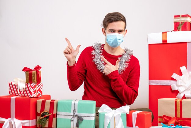 Giovane di vista frontale con il dito della maschera che indica qualcosa che si siede intorno ai regali di natale