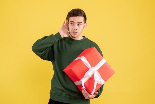 Giovane di vista frontale con il maglione verde che ascolta qualcosa che tiene regalo sul colore giallo