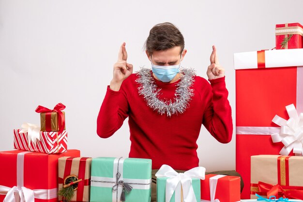 Giovane di vista frontale con la maschera che fa il segno di buona fortuna che si siede intorno ai regali di natale