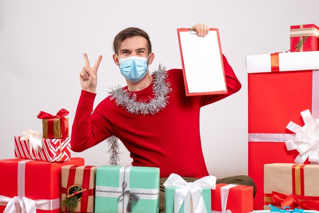 Giovane di vista frontale con la maschera che fa il segno di vittoria che si siede intorno ai regali di natale