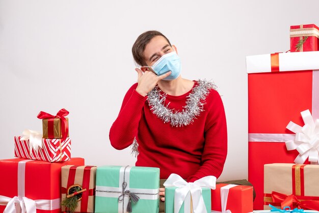 Giovane di vista frontale con la maschera che fa mi chiama segno che si siede intorno ai regali di natale