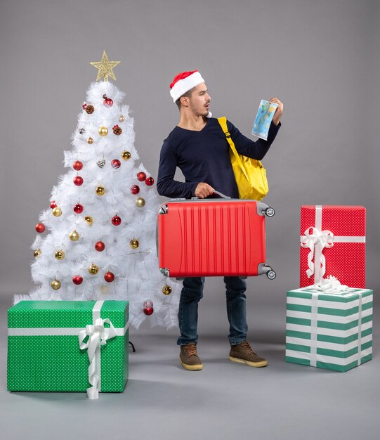 Giovane di vista frontale con lo zaino che tiene la mappa guardando la mappa vicino all'albero di natale su grigio isolato