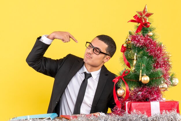 Giovane di vista frontale in un vestito che si siede al tavolo che indica con il dito stesso albero di Natale e regali