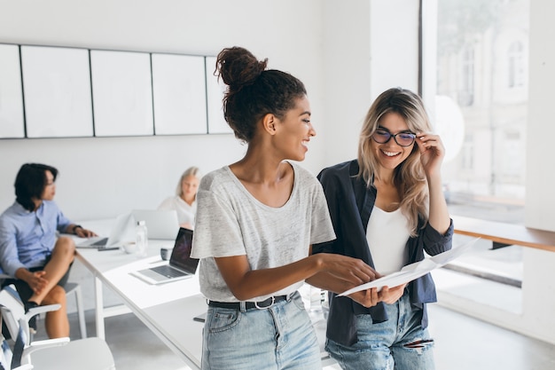 Giovane dirigente femminile spiega la nuova strategia al dipendente biondo in bicchieri e sorridente. Ritratto dell'interno del collettivo multiculturale che lavora al progetto in ufficio e che utilizza computer portatile.