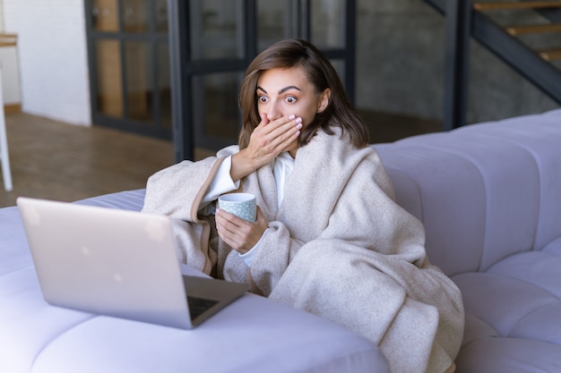 Giovane donna a casa sul divano in inverno sotto un'accogliente coperta con un laptop, guarda scioccata lo schermo