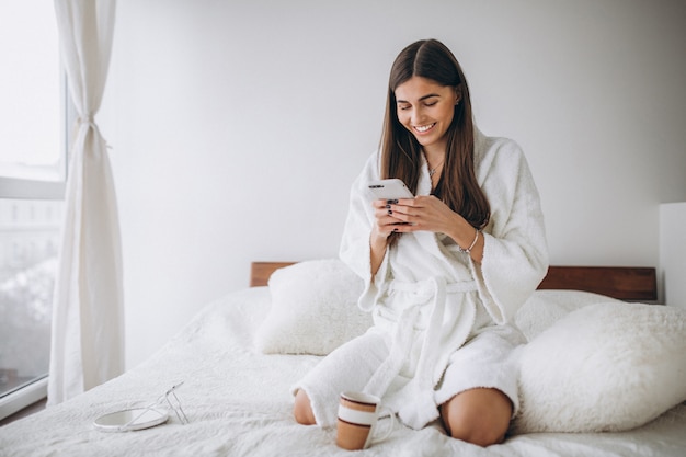 Giovane donna a letto in accappatoio con telefono bere caffè