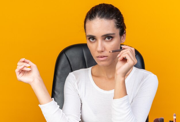 Giovane donna abbastanza caucasica seduta al tavolo con strumenti per il trucco che tengono eyeliner isolato sulla parete arancione con spazio di copia