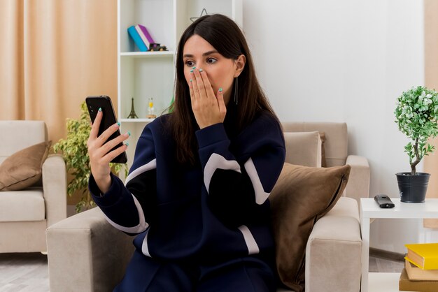 Giovane donna abbastanza caucasica sorpresa che si siede sulla poltrona nel soggiorno progettato che tiene e che esamina il telefono cellulare che tiene la mano sulla bocca