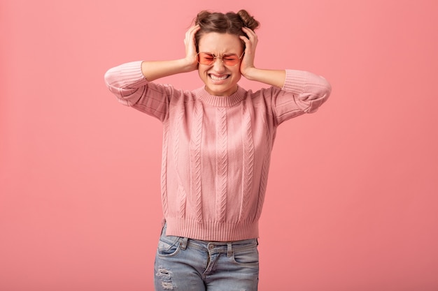 Giovane donna abbastanza divertente che ha un problema, sensazione di mal di testa, stress e tenendo la testa, in maglione rosa e occhiali da sole isolati su sfondo rosa studio