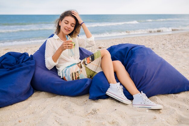 Giovane donna abbastanza elegante che si siede in sacchi di fagioli sulla spiaggia bevendo cocktail mojito, vestito stile estivo, rilassante, gambe in scarpe da ginnastica, aspetto naturale