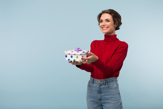 Giovane donna abbastanza sorridente con i capelli corti scuri in maglione rosso che tiene in mano una piccola scatola regalo mentre guarda felicemente da parte su sfondo blu