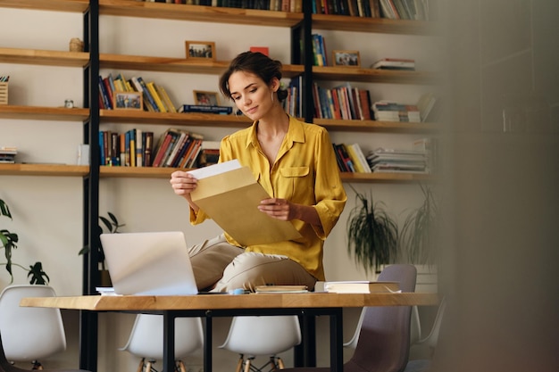 Giovane donna abbastanza sorridente in camicia gialla seduta sulla scrivania con documenti e laptop mentre lavora sognante in un ufficio moderno