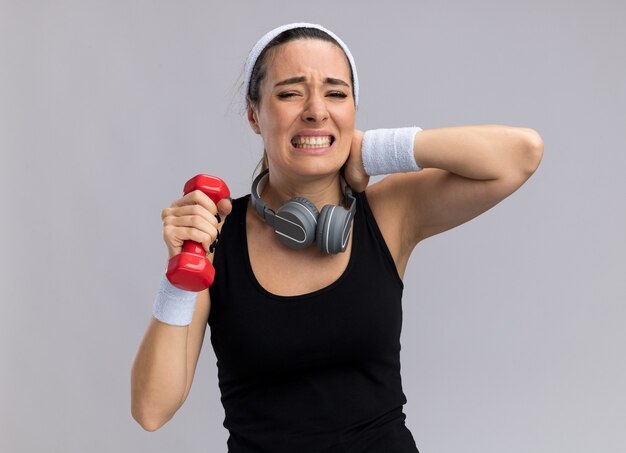 Giovane donna abbastanza sportiva dolorante che indossa fascia e braccialetti che tengono il manubrio con le cuffie intorno al collo tenendo la mano sul collo