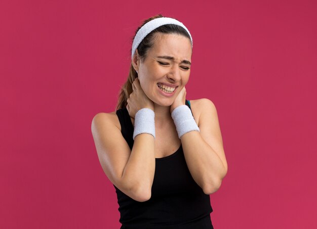 Giovane donna abbastanza sportiva dolorante che indossa fascia e braccialetti che tengono le mani sul collo con gli occhi chiusi