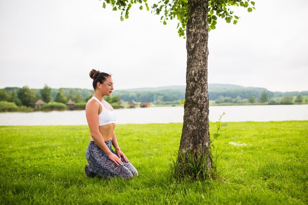 Giovane donna adatta che fa yoga nel parco vicino al lago e all'albero