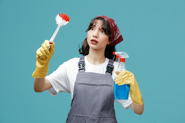 Giovane donna addetta alle pulizie concentrata che indossa bandana uniforme e guanti di gomma che tengono detergente e spazzola guardando il lato finta di pulire qualcosa di isolato su sfondo blu