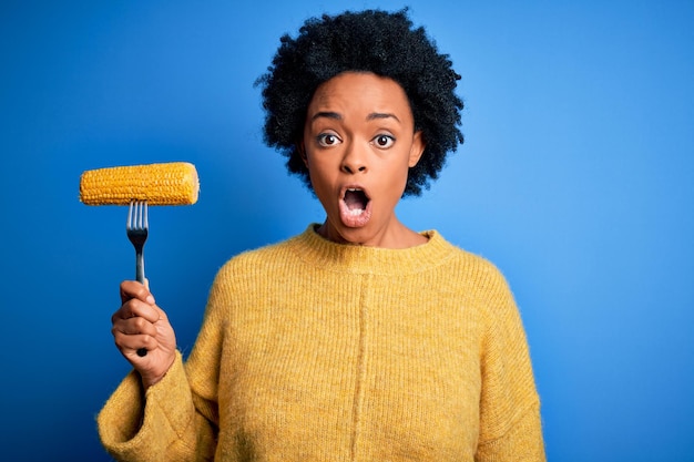 Giovane donna afro-vegetariana afroamericana con i capelli ricci che tiene la forchetta con la pannocchia di mais spaventata dallo shock con una faccia a sorpresa impaurita ed eccitata con l'espressione di paura